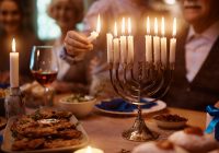 Close up of boy lights candles in menorah while celebrating Hanukkah with his family at dining table.
